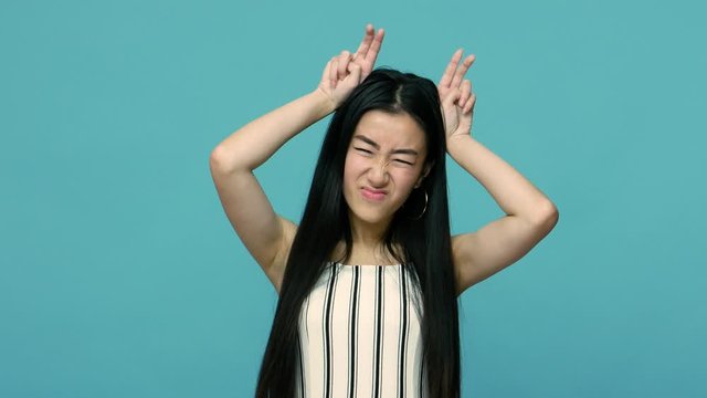 Angry asian woman with long hair threatening with bull horn gesture, looking at camera with hostile bully expression, provoking to conflict, attack. indoor studio shot isolated on blue background
