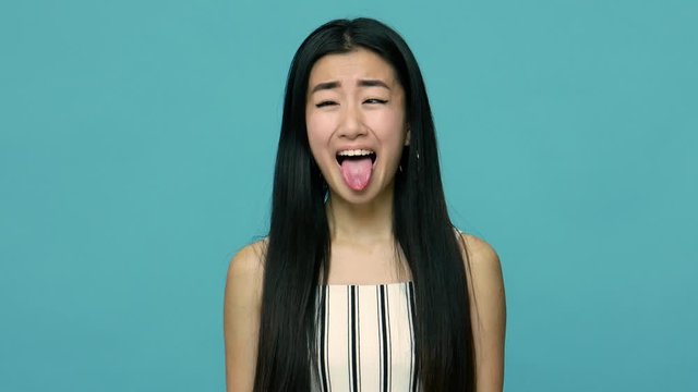 Fooling around. Naughty funny asian woman with long black hair making silly face with crossed eyes and sticking out tongue, looking childish and dumb. indoor studio shot isolated on blue background