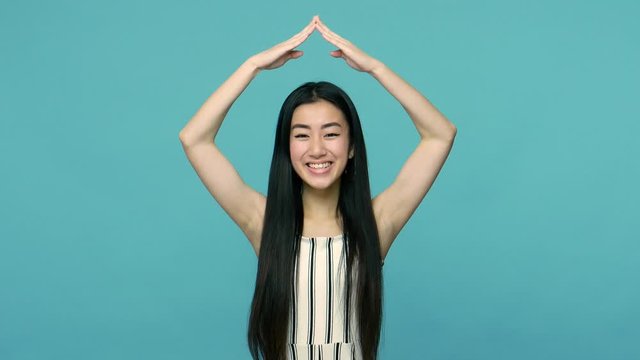 Cheerful tiny asian woman with long straight black hair making roof symbol with hands over head and smiling at camera, feeling safe and protected, house insurance. indoor studio shot, blue background
