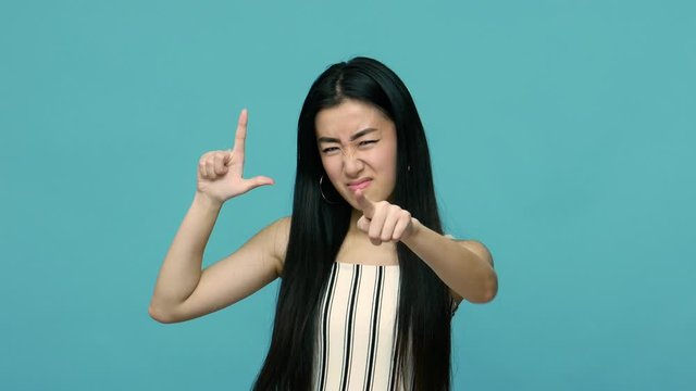 You are loser, your fault! Angry bossy asian woman with long black hair showing loser gesture and pointing at camera, blaming accusing in failure. indoor studio shot isolated on blue background