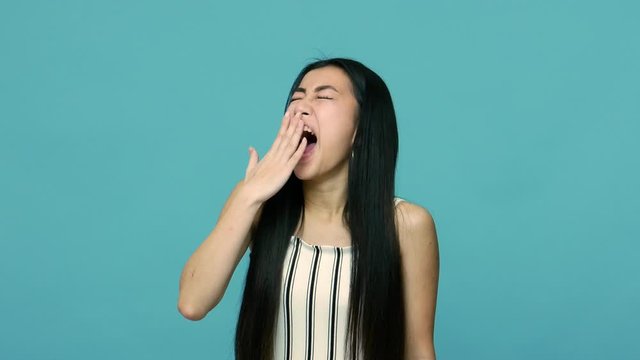 Sleepy overworked asian woman with long black hair yawning and covering mouth with hand, fatigued employee feeling drowsy and lazy, lack of energy. indoor studio shot isolated on blue background