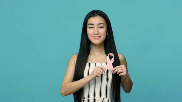 Happy beautiful asian woman with long black hair holding pink ribbon, symbol of breast cancer awareness, female healthcare concept and medical insurance. indoor studio shot isolated on blue background