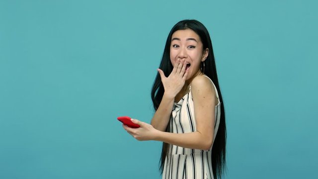 Surprised by mobile app. Happy asian woman with long hair in dress scrolling application using cell phone and looking at camera with amazement, reading shocking message in social media. studio shot