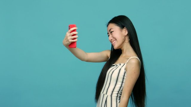 Stunning slim asian woman with long black hair and in dress taking selfie with smartphone, gesturing and making face, glamour girl doing photos on mobile phone. indoor studio shot, blue background
