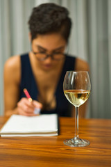 A woman wearing glasses sits at a table drinking white wine and writing in a journal