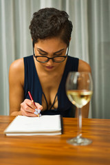 A woman wearing glasses sits at a table drinking white wine and writing in a journal