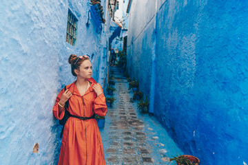 a tourist in a bright long African dress with a belted strap stands in a very narrow street of the blue city of Morocco