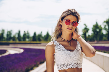 Fototapeta premium girl in sunglasses and a lace topic is standing in the park against the background of flower beds of blooming sage