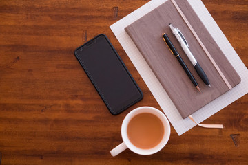 Cup of coffee/tea with black smartphone/cellphone with notebook and pens on a wooden desktop. Working from home, or at a coffee shop. or nice wooden tabletop overhead lay flat shot. Ready for a call.