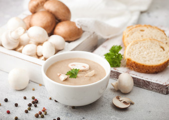White bowl plate of creamy chestnut champignon mushroom soup on white kitchen background and box of raw mushrooms and fresh bread.
