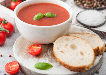 White bowl plate of creamy tomato soup with spoon on light table background with box of raw tomatoes and bread.