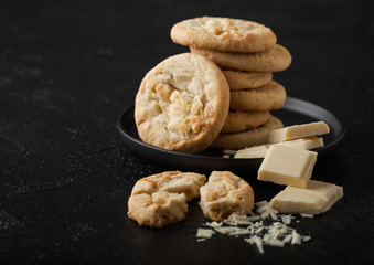 White chocolate biscuit cookies on black ceramic plate with chocolate blocks and curls on dark kitchen table background.