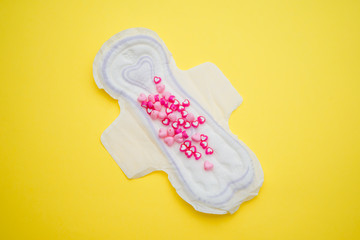 Menstrual pad with heart shape beads on yellow colored background. Woman periods cycle, menstruation frequency. Minimalist still life photography concept
