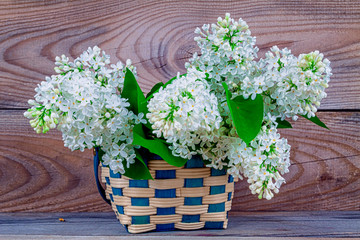 basket with a branch of lilac flower on a wooden background.