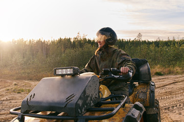 Young beautiful tanned woman dressed in special clothes under disguise wears helmet with glasses rides powerful Quad bike with large studded wheels, girl smiles, looks happy. Against background forest © EverGrump