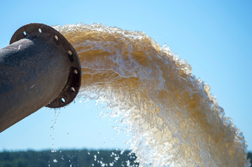 A stream of muddy water bursts out of the pipe against the blue sky and the green forest in the...
