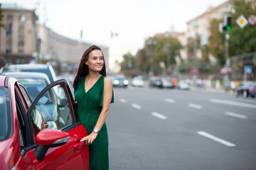 Fashion model wearing green overall posing outdoor next to her red car on the road. Young beautiful brunette caucasian woman driver. Beautiful girl, urban portrait.