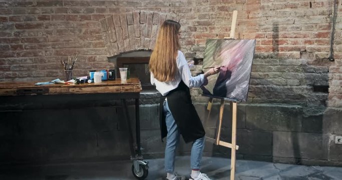 Backside view of talented innovative pretty female artist draws with brush on the canvas. Contemporary young woman painter creating abstract picture. Attractive girl creates modern art painting