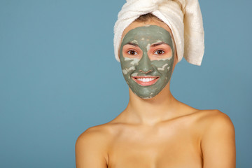 Beautiful cheerful girl applying facial clay mask. Young woman and beauty treatments over blue background. Pampering, youth, anti-aging, body care spa theme. Female face with cosmetics mask