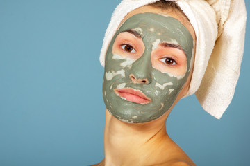 Beautiful cheerful girl applying facial clay mask. Young woman and beauty treatments over blue background. Pampering, youth, anti-aging, body care spa theme. Female face with cosmetics mask