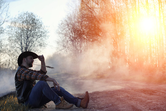 A man is wearing a cowboy hat and a loso in the field. American farmer in a field wearing a jeans hat and with a lasso in the smoke of a fire. A man walks through a burning field