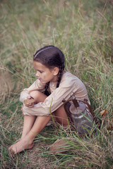 Pretty little girl with long pigtails and beautiful dirty face sitting at summer meadow with vintage paraffin stove and eating stale bread. Orphan, child of war, poverty, hunger, famine