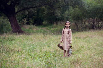 Pretty little girl with long pigtails and beautiful dirty face stands at summer meadow and holding vintage paraffin stove and flask. Orphan, child of war, poverty, hunger, famine, poor destitute kid.