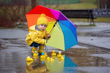 Beautiful funny blonde toddler boy with rubber ducks and colorful umbrella, jumping in puddles and...
