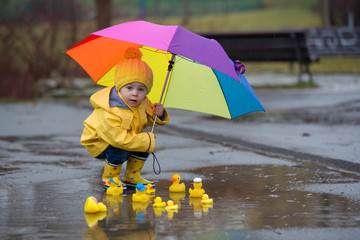 Beautiful funny blonde toddler boy with rubber ducks and colorful umbrella, jumping in puddles and...