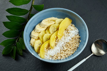 Close up view on Healthy breakfast: oatmeal with coconut, mango, bananas and yellow kiwi in a blue bowl on a black background.
