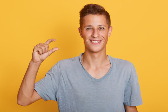 Close up portrait of handsome young man with stylish hairstyle shows tiny little thing. Demonstrates small thing, poaing isolated over yellow background in studio, looking smoling at camera.