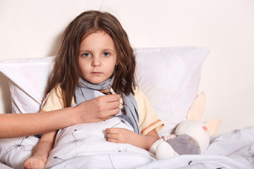 Mother taking temperature of sick daughter. Family takes care of little dark haired girl, female kid looks at camera with sad facial expression, being in bed, lying on white linen. Health care concept