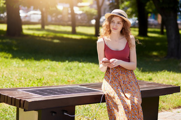 Naklejka premium Photo of attractive woman with foxy hair, wearing hat, floor skirt and maroon t shirt, sitting on bench in city park, charging smart phone via alternative energy. Alternative energy, ecology concept.