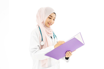 Portrait of smiley beautiful young muslim woman doctor wearing doctor uniform and Hijab and Stethoscope writing in her note book, standing isolated on white background