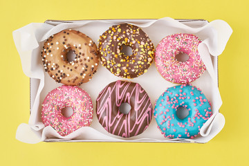 Different types of colorful donats decorated sprinkles and icing in box on a yellow background, top...