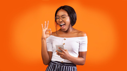 Positive Girl Holding Phone Gesturing Okay And Winking, Orange Background
