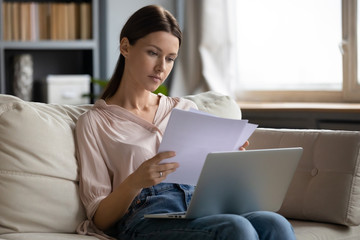 Focused young woman consider paperwork working on laptop