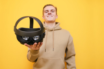 Joyful young man in casual clothes stands on a yellow background with a VR helmet in his hand, looks into the camera and smiles. Focus on helmet in the hands of happy guy. Gaming in virtual reality