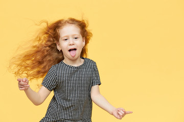 Body language and human emotions. Picture of funny playful Caucasian girl wearing checkered dress pointing fore fingers and sticking out tongue as if teasing you, having fun and fooling around