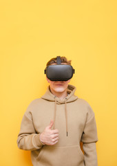 Positive teen guy in virtual reality helmet and casual clothes isolated on yellow background, showing thumbs up and smiling.Young man wishing to use VR technology, wears a helmet.Student recommends VR
