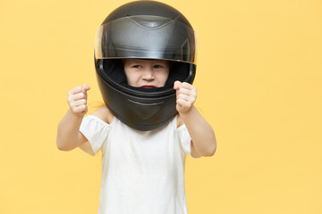 Close up shot of confident furious little girl wearing protective head equipment while driving sports car keeping hands in front of her as if holding them on steering wheel. Adrenaline and extreme
