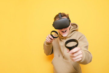 Cheerful guy stands with controllers in his hands and VR helmet on his head against a yellow background and plays video games with a funny face. Young gamer playing VR games with helmet and smiling.