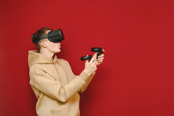 Portrait of surprised VR gamer with virtual reality helmet on his head,playing video games on red background,looking away at copy space.Guy plays VR games with controllers in his hands and a VR helmet