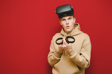 Emotional gamer in VR on head expressively plays games on controllers, isolated on red background.Surprised gamer plays VR games and looks in camera. Copy space