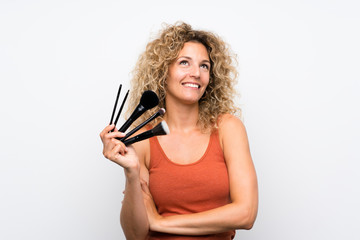 Young blonde woman with curly hair holding a lot of makeup brush looking up while smiling