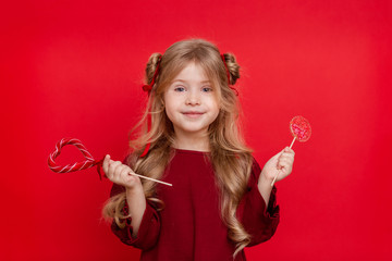 Portrait of a cheerful dreamy little girl with a heart shaped candy in her hands isolated on a red...