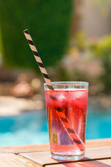 Red cocktail with ice with a straw on the background of the pool. vertical photo