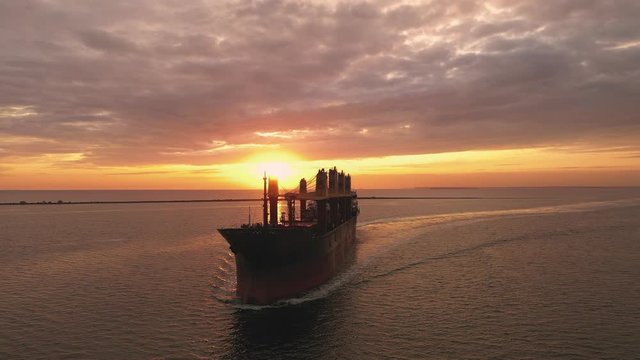 Slow-motion cargo bulker ship, bulk carrier with deck cranes sailing in sunset, front view merchant vessel transporting grains, crops via sea, arriving seaport evening, shipping wheat, maize harbour