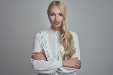 Young and beautiful long-haired blond woman in white blouse standing with her arms folded and looking at camera with a smirk. Front half-length portrait against grey background