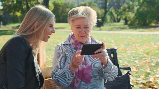 Elder grandmother showing her granddaughter photos on smartphone. They sitting on bench and talking in autumn park.